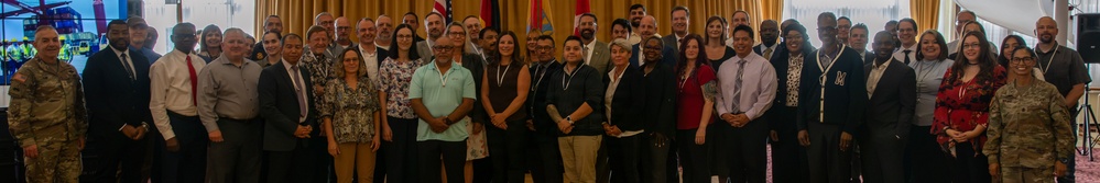A large group of approximately 50-60 individuals are posed together for a formal group photograph in what appears to be a conference or event venue. The group includes both military personnel in uniform and civilians in business/professional attire. Multiple national flags are visible in the background, suggesting an international or multinational gathering.