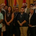 A large group of approximately 50-60 individuals are posed together for a formal group photograph in what appears to be a conference or event venue. The group includes both military personnel in uniform and civilians in business/professional attire. Multiple national flags are visible in the background, suggesting an international or multinational gathering.