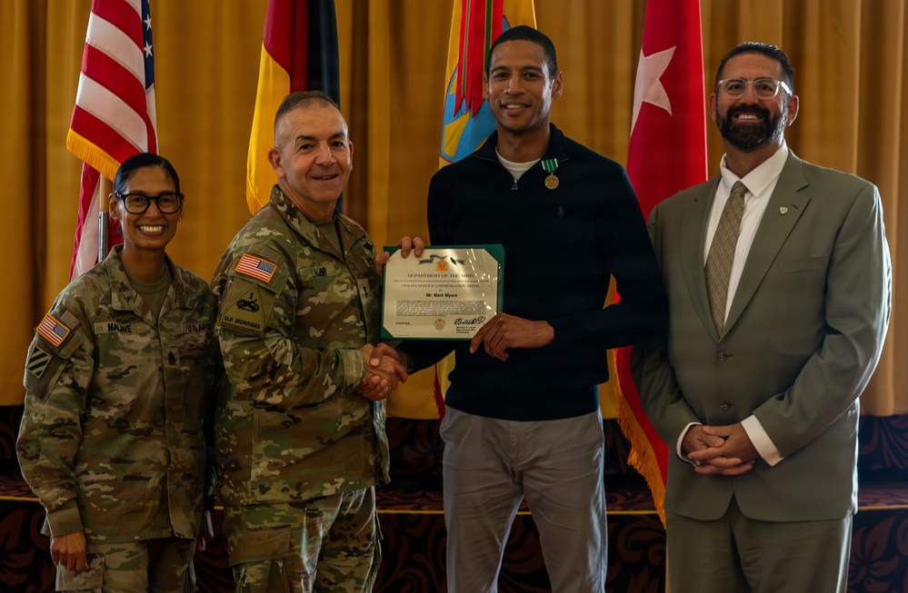 A formal military award ceremony is taking place, with a U.S. Army officer presenting a Department of the Army Civilian Service Commendation certificate to a civilian recipient named 'Mr. Mark Myers,' as visible on the certificate. Four individuals are posed together: two in U.S. Army combat uniforms, one civilian recipient wearing a commendation medal, and one civilian in a suit. Multiple flags are displayed in the background, including the U.S. flag and what appears to be a German flag.