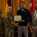 A formal military award ceremony is taking place, with a U.S. Army officer presenting a Department of the Army Civilian Service Commendation certificate to a civilian recipient named 'Mr. Mark Myers,' as visible on the certificate. Four individuals are posed together: two in U.S. Army combat uniforms, one civilian recipient wearing a commendation medal, and one civilian in a suit. Multiple flags are displayed in the background, including the U.S. flag and what appears to be a German flag.