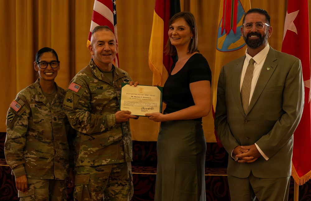 A formal Department of the Army certificate presentation ceremony is taking place indoors. Two military personnel in Army Combat Uniforms (ACUs) are presenting a certificate to a civilian woman, while a civilian man in a suit stands to the right. Multiple flags, including the U.S. flag and what appears to be unit/organizational flags, are displayed in the background.
