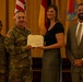 A formal Department of the Army certificate presentation ceremony is taking place indoors. Two military personnel in Army Combat Uniforms (ACUs) are presenting a certificate to a civilian woman, while a civilian man in a suit stands to the right. Multiple flags, including the U.S. flag and what appears to be unit/organizational flags, are displayed in the background.