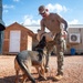 A military or law enforcement handler in camouflage pants and a tan shirt is working with a German Shepherd dog, using a burlap training sleeve or reward toy during what appears to be a K9 training exercise. The setting is an outdoor compound with wooden structures and shipping containers, consistent with a forward operating base or training facility. A second person in dark clothing is partially visible in the foreground.