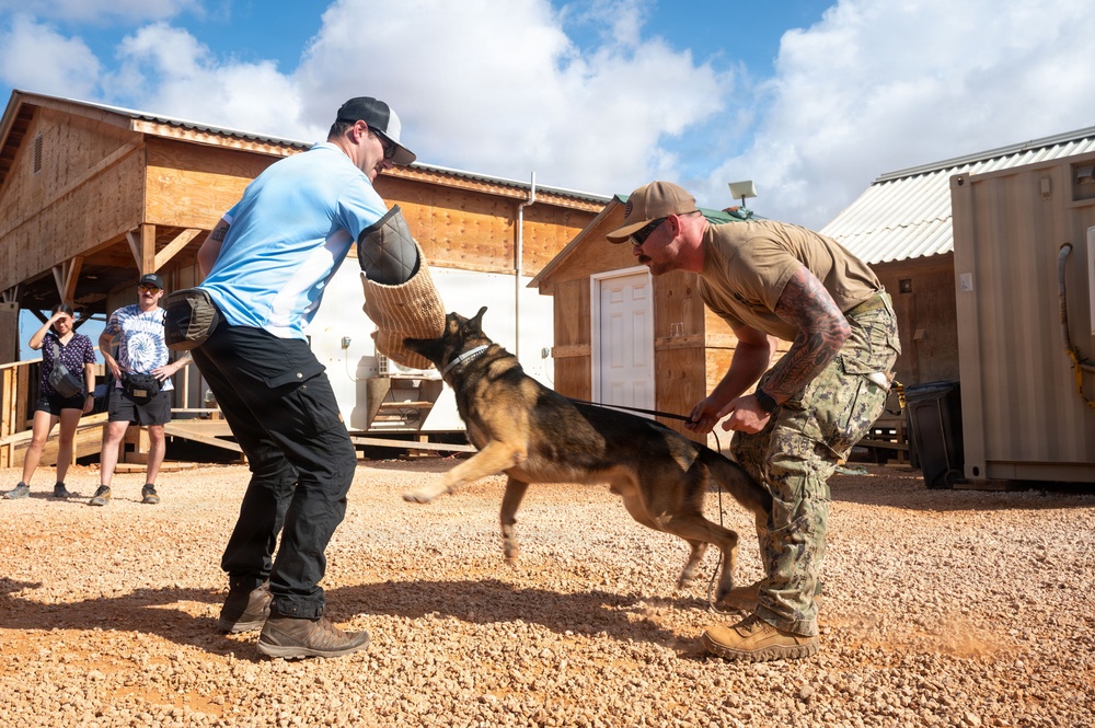A military working dog (MWD) training demonstration is taking place on a gravel surface near wooden structures. A handler in camouflage uniform controls the dog on a leash while the dog bites a protective sleeve worn by a decoy in civilian clothing. Observers watch from the background near a wooden building.