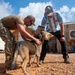 Two men are interacting with a military working dog, likely a German Shepherd, in an outdoor compound area with gravel ground and shipping container structures. One man is wearing military camouflage uniform while the other is in civilian clothing including an Adidas Argentina soccer jersey. The activity appears to be a training exercise or reward session involving a burlap/jute training bite sleeve.