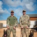 Two military service members pose with their military working dogs (MWDs) at what appears to be a forward operating base. The person on the left wears U.S. Navy NWU Type III (digital camouflage) uniform, while the person on the right wears U.S. Air Force OCP (Operational Camouflage Pattern) uniform with visible 'U.S. AIR FORCE' tape and a name tape reading 'ZEIGT'. Each handler stands beside a Belgian Malinois or German Shepherd MWD on a leash.