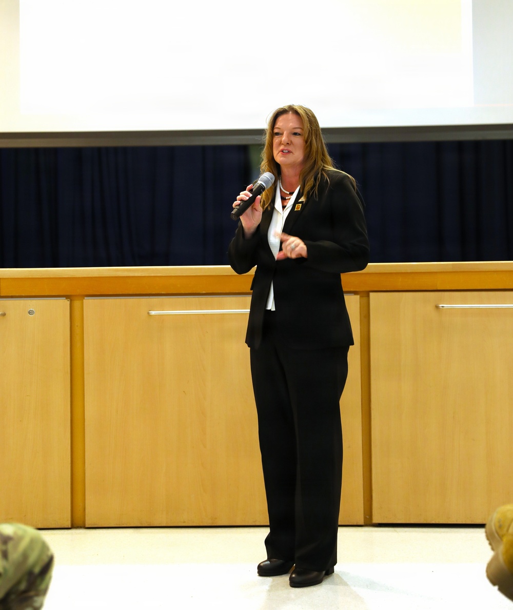 A woman in a black business suit is standing and speaking into a handheld microphone in what appears to be a formal presentation or briefing setting. She is positioned in front of a projection screen and wooden paneled backdrop, addressing an audience. At least one person in military camouflage uniform is partially visible in the foreground.