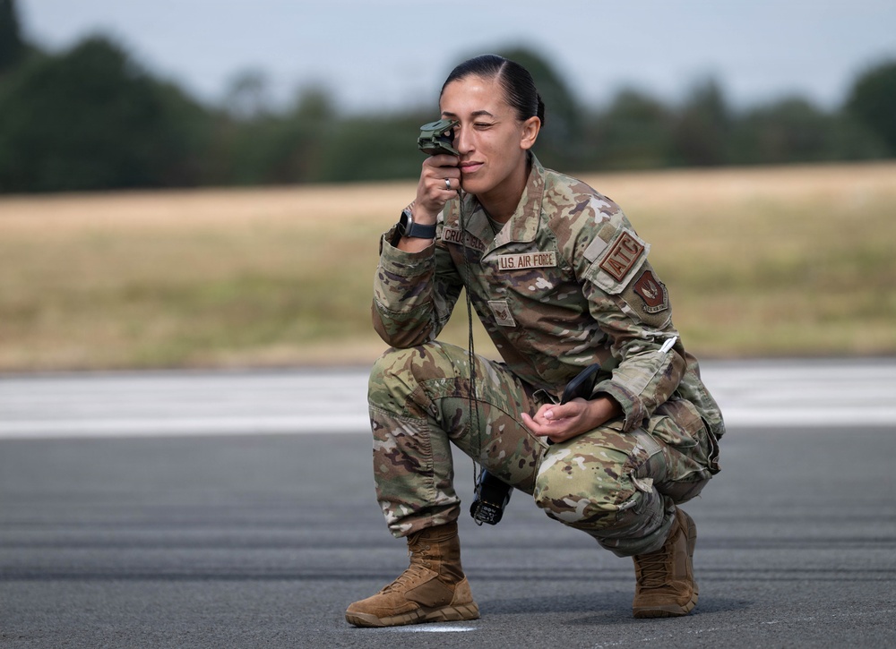 A U.S. Air Force service member in OCP (Operational Camouflage Pattern) uniform is crouched on what appears to be a runway or tarmac, using a handheld optical device held up to one eye. The name tape reads 'U.S. AIR FORCE' and a partial last name beginning with 'CRUZ-' is visible, along with an 'ATC' (Air Traffic Control) patch. The individual appears to be conducting airfield or runway assessment operations.