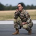 A U.S. Air Force service member in OCP (Operational Camouflage Pattern) uniform is crouched on what appears to be a runway or tarmac, using a handheld optical device held up to one eye. The name tape reads 'U.S. AIR FORCE' and a partial last name beginning with 'CRUZ-' is visible, along with an 'ATC' (Air Traffic Control) patch. The individual appears to be conducting airfield or runway assessment operations.