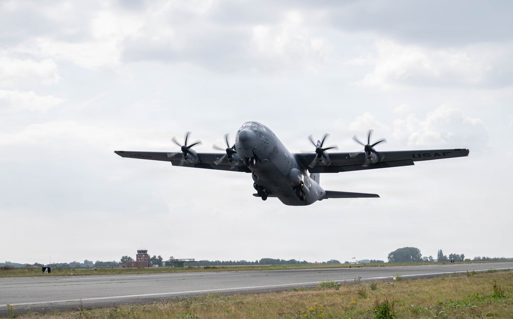A USAF C-130J Super Hercules military transport aircraft is captured at the moment of takeoff from a runway, with all four turboprop engines at full power and propellers visibly spinning. The aircraft is painted in overall gray livery with 'USAF' markings visible on the tail section. A control tower and tree line are visible in the background under an overcast sky.