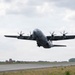 A USAF C-130J Super Hercules military transport aircraft is captured at the moment of takeoff from a runway, with all four turboprop engines at full power and propellers visibly spinning. The aircraft is painted in overall gray livery with 'USAF' markings visible on the tail section. A control tower and tree line are visible in the background under an overcast sky.