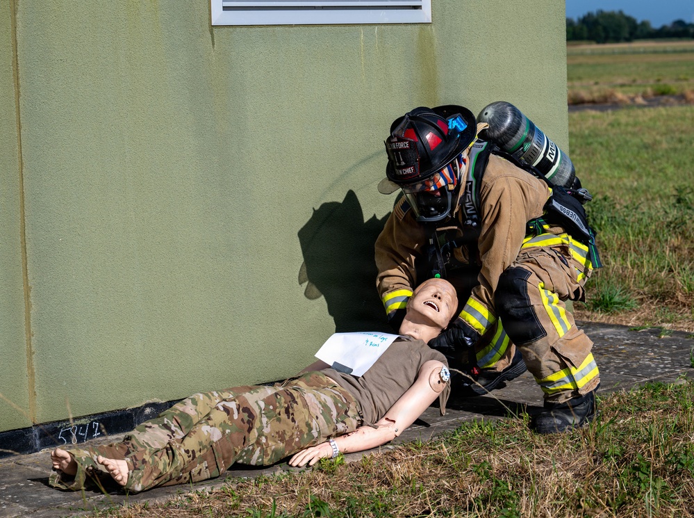 A firefighter in full turnout gear and SCBA (self-contained breathing apparatus) is attending to a training mannequin dressed in military camouflage uniform, lying on the ground beside a green building exterior. The scene depicts a military emergency response training exercise, as indicated by the realistic mannequin used as a simulated casualty. The setting appears to be an outdoor training area on a military installation.