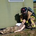 A firefighter in full turnout gear and SCBA (self-contained breathing apparatus) is attending to a training mannequin dressed in military camouflage uniform, lying on the ground beside a green building exterior. The scene depicts a military emergency response training exercise, as indicated by the realistic mannequin used as a simulated casualty. The setting appears to be an outdoor training area on a military installation.