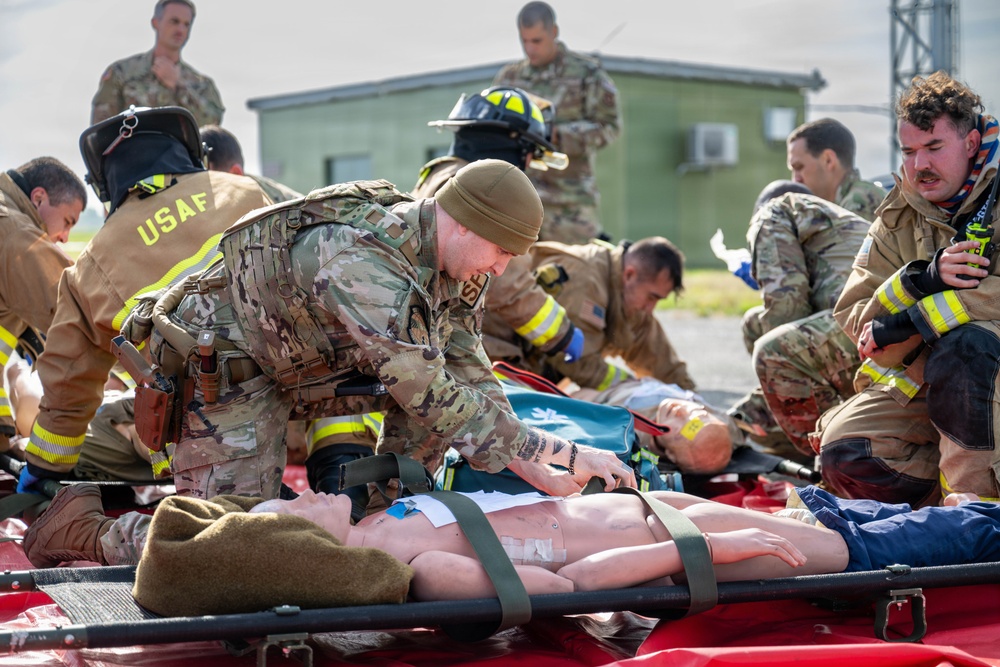 A military mass casualty training exercise is underway outdoors, involving USAF personnel in both combat uniforms and firefighter gear. Multiple simulated casualties (mannequins and role-players) are laid out on stretchers and red mats while responders practice triage and medical treatment procedures. The scene depicts coordinated interoperability between military medics and USAF firefighters.