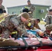 A military mass casualty training exercise is underway outdoors, involving USAF personnel in both combat uniforms and firefighter gear. Multiple simulated casualties (mannequins and role-players) are laid out on stretchers and red mats while responders practice triage and medical treatment procedures. The scene depicts coordinated interoperability between military medics and USAF firefighters.
