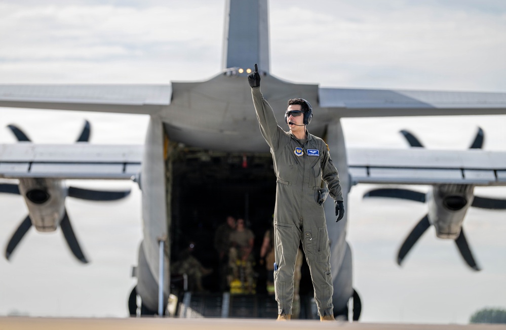 A military aircrew member in an olive/sage green flight suit stands in front of a large military transport aircraft, raising one finger upward in a marshaling or signaling gesture. The aircraft appears to be a C-130 Hercules or similar turboprop transport, with its rear cargo ramp open and personnel visible inside. The scene takes place on an active flight line or tarmac.