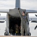 A military aircrew member in an olive/sage green flight suit stands in front of a large military transport aircraft, raising one finger upward in a marshaling or signaling gesture. The aircraft appears to be a C-130 Hercules or similar turboprop transport, with its rear cargo ramp open and personnel visible inside. The scene takes place on an active flight line or tarmac.