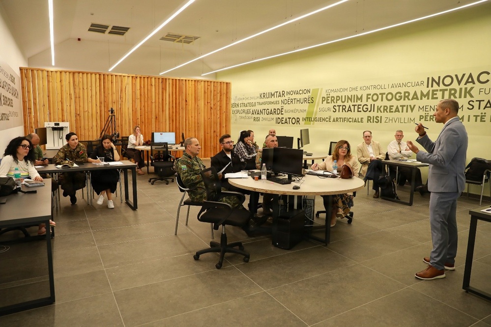 A professional briefing or training session is taking place in a modern innovation-themed room, with a presenter in a gray suit addressing a mixed audience of military personnel in camouflage uniforms and civilians. The wall features Albanian-language text including words like 'INOVACION', 'FOTOGRAMETRIK', 'STRATEGJI', and 'INTELIGJENCË ARTIFICIALE', suggesting a technology or innovation-focused military or government facility. The setting appears to be in Albania based on the visible language.