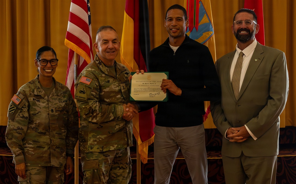 A formal U.S. Army award ceremony showing four individuals posing for a photo. A senior military officer in Army Combat Uniform is presenting a Department of the Army certificate to a civilian male while two others stand alongside them. The setting appears to be an indoor venue with U.S. and other military/national flags displayed in the background.