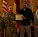 A formal U.S. Army award ceremony showing four individuals posing for a photo. A senior military officer in Army Combat Uniform is presenting a Department of the Army certificate to a civilian male while two others stand alongside them. The setting appears to be an indoor venue with U.S. and other military/national flags displayed in the background.