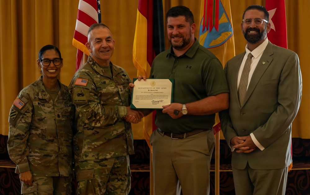 A formal military award ceremony showing four individuals posing for a photo. A uniformed Army soldier is presenting a Department of the Army certificate to a civilian male, while two other individuals stand alongside them. The setting appears to be an official military venue with multiple flags displayed in the background.