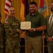 A formal military award ceremony showing four individuals posing for a photo. A uniformed Army soldier is presenting a Department of the Army certificate to a civilian male, while two other individuals stand alongside them. The setting appears to be an official military venue with multiple flags displayed in the background.