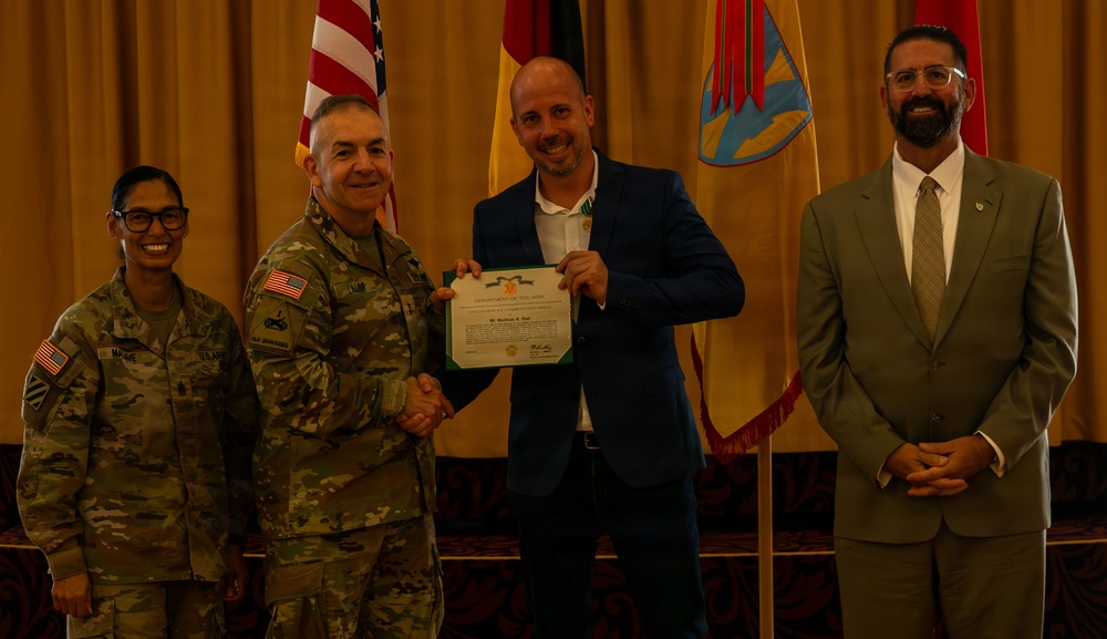A formal military award ceremony is taking place in what appears to be a banquet or conference room. A military officer in Army Combat Uniform is shaking hands with a civilian man in a blue blazer who is holding a Department of the Army certificate. Two additional individuals — a female soldier and a civilian man in a suit — stand alongside them as witnesses.