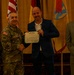 A formal military award ceremony is taking place in what appears to be a banquet or conference room. A military officer in Army Combat Uniform is shaking hands with a civilian man in a blue blazer who is holding a Department of the Army certificate. Two additional individuals — a female soldier and a civilian man in a suit — stand alongside them as witnesses.