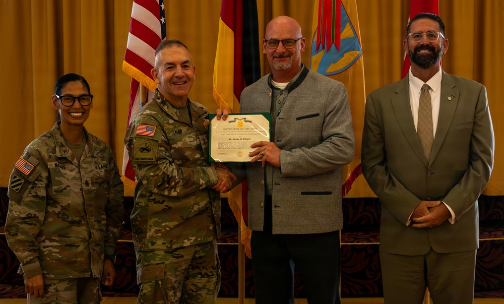 A formal military award ceremony is taking place in what appears to be a banquet or conference hall. A U.S. Army officer in camouflage uniform is presenting a Department of the Army certificate to a civilian man, while two other individuals — a female soldier and a civilian man in a suit — stand alongside them. Multiple flags, including the U.S. flag and unit/state flags, are displayed in the background.
