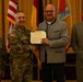 A formal military award ceremony is taking place in what appears to be a banquet or conference hall. A U.S. Army officer in camouflage uniform is presenting a Department of the Army certificate to a civilian man, while two other individuals — a female soldier and a civilian man in a suit — stand alongside them. Multiple flags, including the U.S. flag and unit/state flags, are displayed in the background.