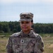 A U.S. Army soldier named Baldwin stands at attention in an outdoor field setting, wearing the standard Operational Camouflage Pattern (OCP) uniform. The soldier is posing for what appears to be an official or documentary-style portrait photograph. A treeline and open grassy terrain are visible in the background.