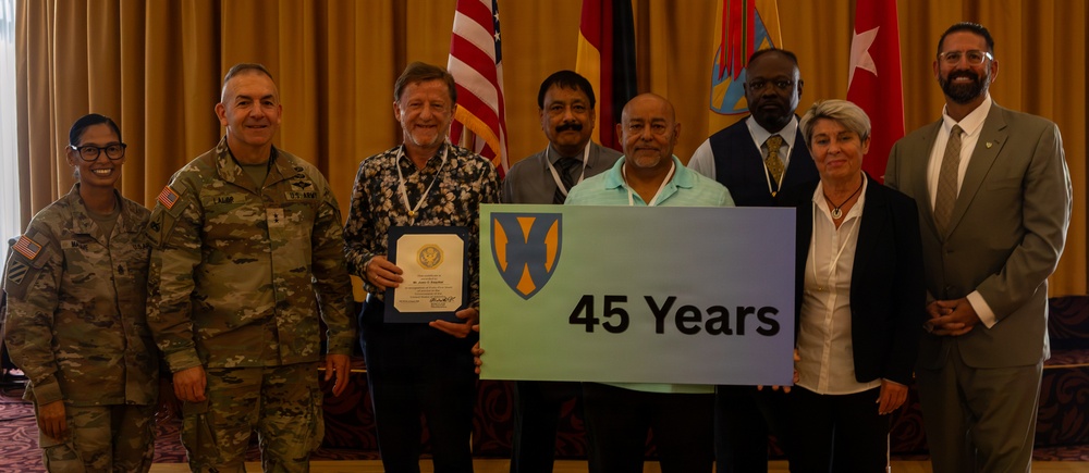 A group of eight people pose for a formal recognition photo at what appears to be a military ceremony or awards event. Two individuals in the center hold a sign displaying '45 Years' featuring a military unit insignia, while one person holds a certificate or award. The group includes two uniformed U.S. Army personnel and several civilians.