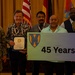 A group of eight people pose for a formal recognition photo at what appears to be a military ceremony or awards event. Two individuals in the center hold a sign displaying '45 Years' featuring a military unit insignia, while one person holds a certificate or award. The group includes two uniformed U.S. Army personnel and several civilians.