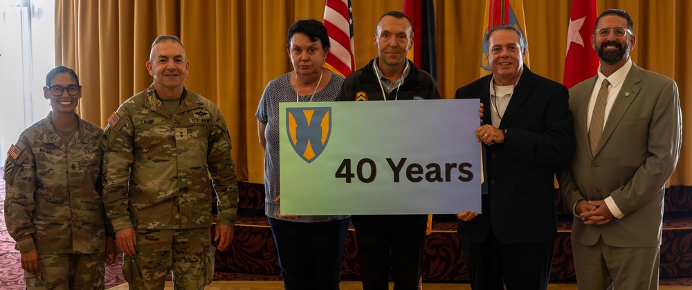 A group of six individuals pose for a formal photo at what appears to be a military recognition ceremony. Two individuals in U.S. Army combat uniforms stand alongside four civilians, with two people holding a sign displaying a military unit insignia and '40 Years.' The setting includes multiple flags in the background, suggesting an official military or government venue.
