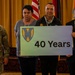 A group of six individuals pose for a formal photo at what appears to be a military recognition ceremony. Two individuals in U.S. Army combat uniforms stand alongside four civilians, with two people holding a sign displaying a military unit insignia and '40 Years.' The setting includes multiple flags in the background, suggesting an official military or government venue.