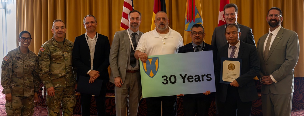 A group of nine individuals pose for a formal commemorative photo, with one person holding a sign reading '30 Years' featuring a military unit insignia. Two individuals are in U.S. Army military uniforms while the others are in business attire. The setting appears to be an indoor event venue with multiple national and organizational flags displayed in the background.