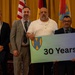 A group of nine individuals pose for a formal commemorative photo, with one person holding a sign reading '30 Years' featuring a military unit insignia. Two individuals are in U.S. Army military uniforms while the others are in business attire. The setting appears to be an indoor event venue with multiple national and organizational flags displayed in the background.