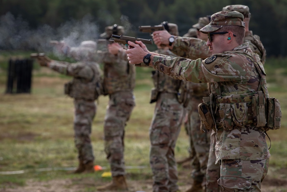 A line of military personnel in OCP (Operational Camouflage Pattern) uniforms are conducting a pistol qualification or training exercise on an outdoor firing range. Multiple soldiers are simultaneously firing semi-automatic pistols downrange, with visible muzzle smoke present on at least one weapon. The foreground soldier is in sharp focus while others recede into bokeh depth-of-field blur.