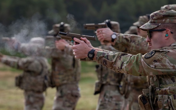 A line of military personnel in OCP (Operational Camouflage Pattern) uniforms are conducting a pistol qualification or training exercise on an outdoor firing range. Multiple soldiers are simultaneously firing semi-automatic pistols downrange, with visible muzzle smoke present on at least one weapon. The foreground soldier is in sharp focus while others recede into bokeh depth-of-field blur.