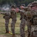 A line of military personnel in OCP (Operational Camouflage Pattern) uniforms are conducting a pistol qualification or training exercise on an outdoor firing range. Multiple soldiers are simultaneously firing semi-automatic pistols downrange, with visible muzzle smoke present on at least one weapon. The foreground soldier is in sharp focus while others recede into bokeh depth-of-field blur.