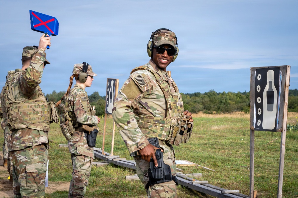 Military personnel in MultiCam/OCP camouflage uniforms are participating in a firearms qualification or training exercise at an outdoor shooting range. The soldiers are equipped with tactical gear including body armor, holstered pistols, and hearing protection. Shooting targets are visible in the foreground, and one soldier is holding up a blue scoring/signaling paddle with a red X.