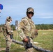 Military personnel in MultiCam/OCP camouflage uniforms are participating in a firearms qualification or training exercise at an outdoor shooting range. The soldiers are equipped with tactical gear including body armor, holstered pistols, and hearing protection. Shooting targets are visible in the foreground, and one soldier is holding up a blue scoring/signaling paddle with a red X.