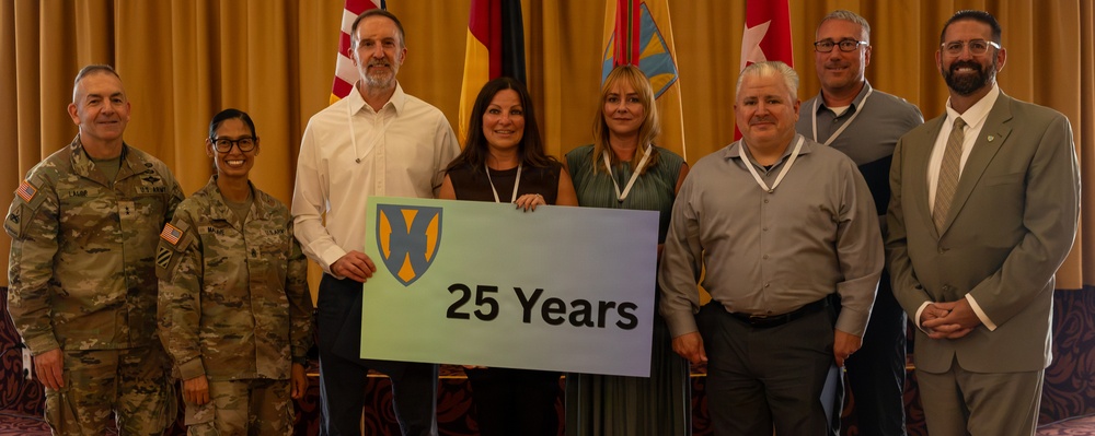 A group of eight people pose for a formal photo at what appears to be a military or government ceremony. Two individuals are wearing U.S. Army military uniforms, while the others are in civilian attire. The group is holding a sign displaying a military unit insignia and the text '25 Years,' commemorating a 25th anniversary milestone.