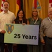 A group of eight people pose for a formal photo at what appears to be a military or government ceremony. Two individuals are wearing U.S. Army military uniforms, while the others are in civilian attire. The group is holding a sign displaying a military unit insignia and the text '25 Years,' commemorating a 25th anniversary milestone.
