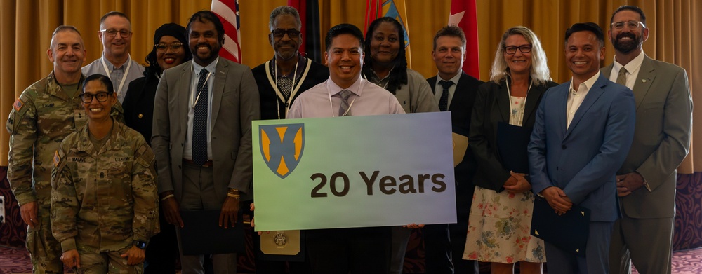 A group of approximately 13 individuals poses for a formal commemorative photo, with one person holding a sign reading '20 Years' featuring a shield emblem. The group includes both military personnel in uniform and civilians in business attire. The setting appears to be an indoor event venue with flags visible in the background.