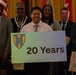 A group of approximately 13 individuals poses for a formal commemorative photo, with one person holding a sign reading '20 Years' featuring a shield emblem. The group includes both military personnel in uniform and civilians in business attire. The setting appears to be an indoor event venue with flags visible in the background.