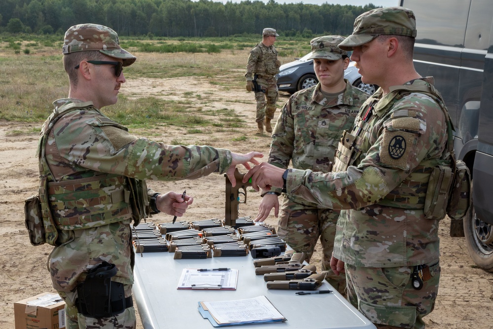U.S. Army soldiers in OCP (Operational Camouflage Pattern) uniforms are conducting a weapons issue or inspection at an outdoor range or training area. A table is laid out with multiple pistols and magazines, and one soldier appears to be handing or receiving a firearm while another observes. The setting appears to be a sandy, open field with tree lines in the background.
