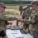 U.S. Army soldiers in OCP (Operational Camouflage Pattern) uniforms are conducting a weapons issue or inspection at an outdoor range or training area. A table is laid out with multiple pistols and magazines, and one soldier appears to be handing or receiving a firearm while another observes. The setting appears to be a sandy, open field with tree lines in the background.