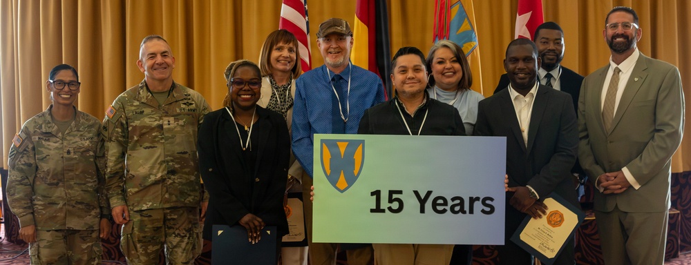 A group of approximately eleven people pose together at what appears to be a formal recognition or awards ceremony. Two individuals in the center are holding a sign displaying a shield logo and the text '15 Years,' suggesting a service anniversary or milestone celebration. The group includes two U.S. Army personnel in military uniforms and several civilians in business attire.