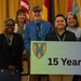 A group of approximately eleven people pose together at what appears to be a formal recognition or awards ceremony. Two individuals in the center are holding a sign displaying a shield logo and the text '15 Years,' suggesting a service anniversary or milestone celebration. The group includes two U.S. Army personnel in military uniforms and several civilians in business attire.